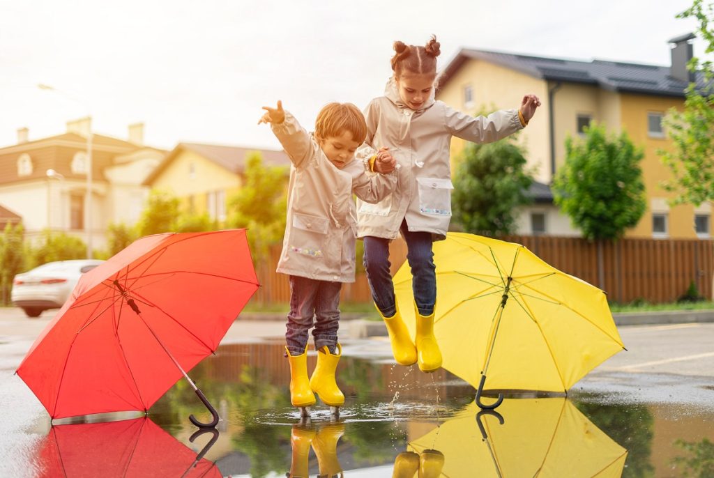 Gyermekek ugrálnak az esőben, gumicsizmában és esőkabátban esernyők mellett.